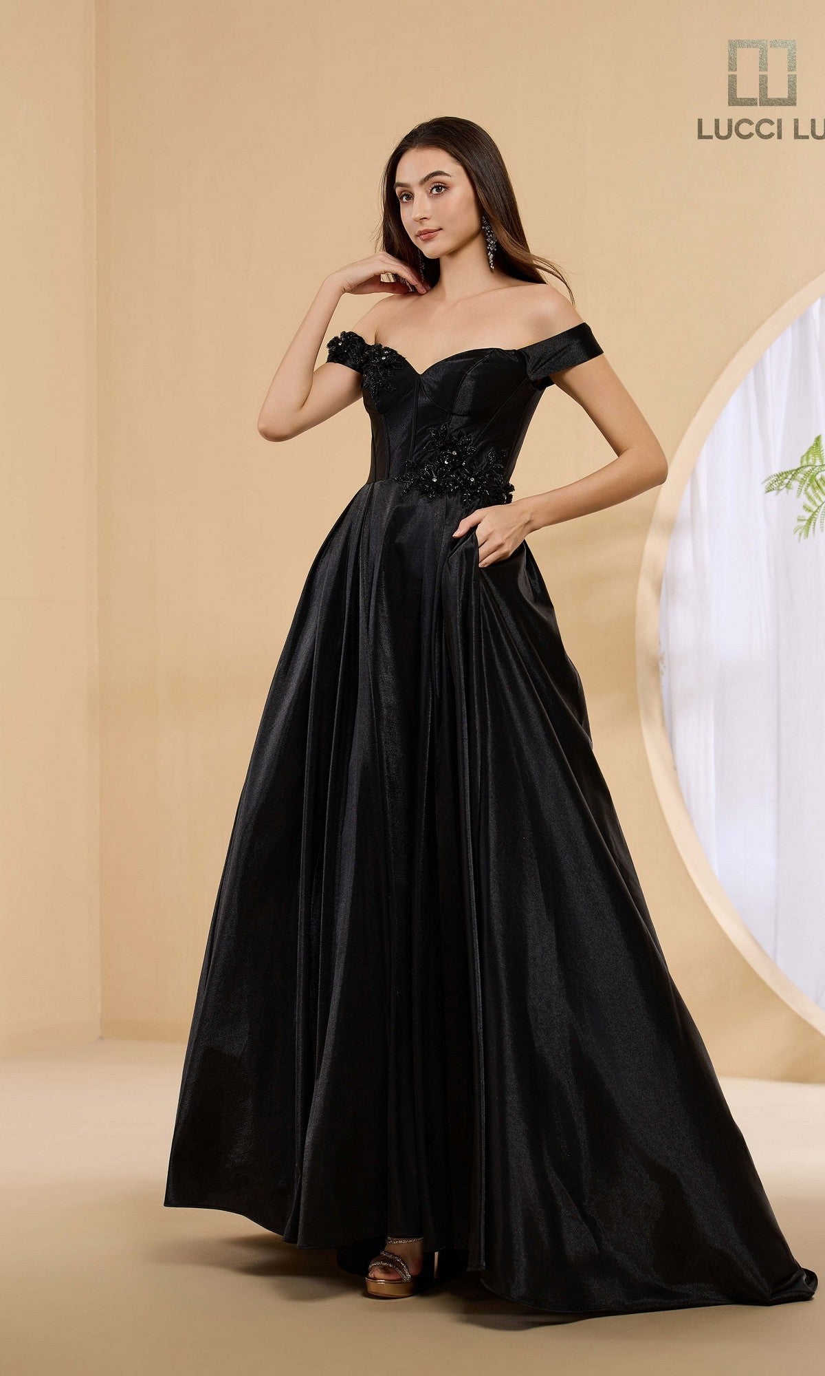 A woman stands against a neutral background wearing the Lucci Lu 1472, a long black prom dress featuring an off-the-shoulder design and floral detailing on the bodice.